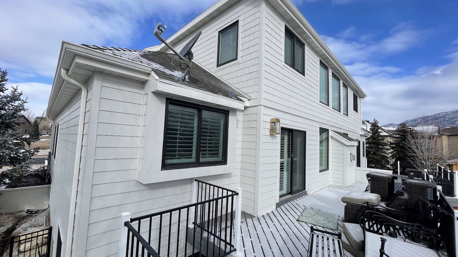 Two-story white house with black trim and durable vinyl siding, a satellite dish on the roof, and a deck furnished with outdoor seating. Snow covers parts of the deck and surrounding ground. Pine trees and mountains are visible, capturing the classic charm of Utah homes. Two-story white house with black trim and durable vinyl siding, a satellite dish on the roof, and a deck furnished with outdoor seating. Snow covers parts of the deck and surrounding ground. Pine trees and mountains are visible, capturing the classic charm of Utah homes.