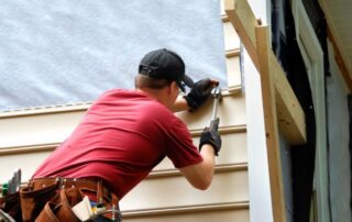 A construction worker in a red shirt and black cap installs beige siding on a Utah new construction home using a hammer.