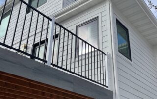 Modern white two-story house exterior finished with James Hardie siding, featuring black-trimmed windows and a sleek black metal balcony railing, viewed from below. Sunlight reflects off a window, highlighting the clean lines often seen in contemporary Utah homes.