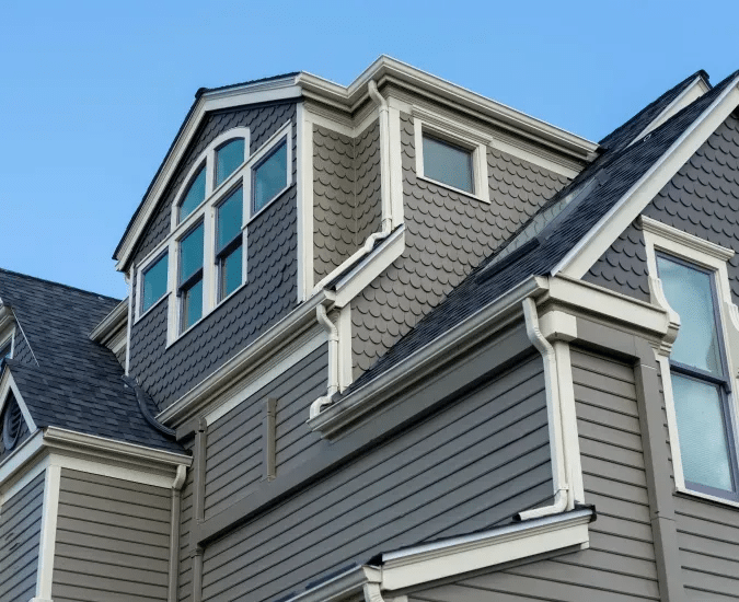 Close-up view of the exterior of a multi-story house featuring remodeled gray siding, white trim, and multiple windows, set against a clear blue sky. A split view of a home interior shows a finished hallway with wooden flooring and staircase on the left, and an active remodeling project underway on the right.