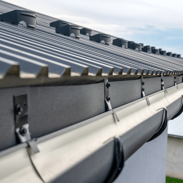 Close-up view of a building’s corrugated metal roof with drip edge, aluminum gutter installation, and downspout attachments against a blue Utah sky. Close-up view of a building’s corrugated metal roof with drip edge, aluminum gutter installation, and downspout attachments against a blue Utah sky.