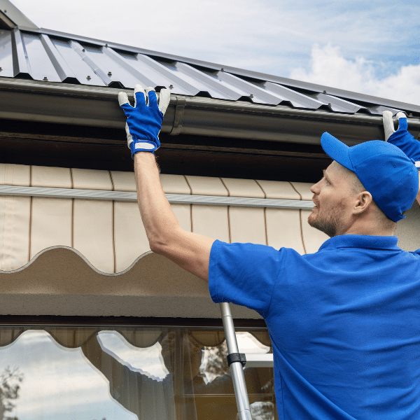 A worker in blue gloves and shirt stands on a ladder, inspecting or repairing an aluminum gutter and metal roof on a house in Salt Lake City. A worker in blue gloves and shirt stands on a ladder, inspecting or repairing an aluminum gutter and metal roof on a house in Salt Lake City.
