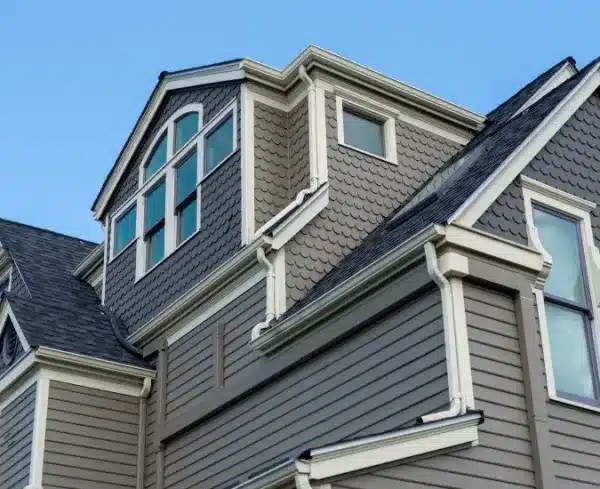 Close-up view of a grey house exterior with decorative siding, white trim, multiple windows, and a gable roof under a clear blue sky. Close-up view of a grey house exterior with decorative siding, white trim, multiple windows, and a gable roof under a clear blue sky.