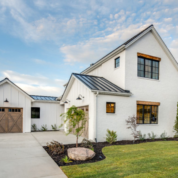 Modern farmhouse in Salt Lake City with white siding, black metal roof, large windows, wooden garage doors, landscaped front yard, Hard Coat Traditional Stucco accents, and concrete driveway under a partly cloudy sky. Modern farmhouse in Salt Lake City with white siding, black metal roof, large windows, wooden garage doors, landscaped front yard, Hard Coat Traditional Stucco accents, and concrete driveway under a partly cloudy sky.