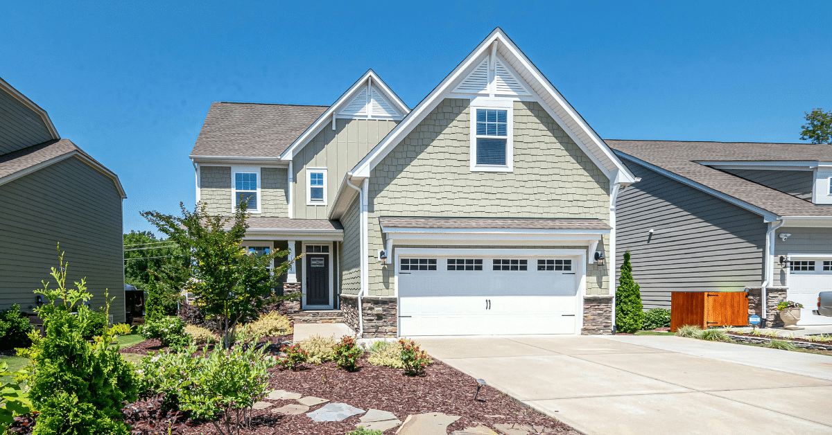 Two-story suburban house with light green siding, white trim, and a double garage—showcasing expert siding installation Utah—surrounded by a landscaped yard and driveway under a clear blue sky.