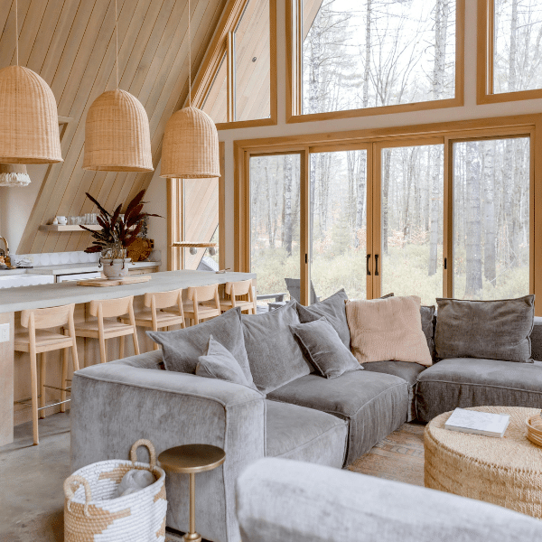 Modern living room with a grey sectional sofa, woven pendant lights, wood accents, and large wood windows offering a view of trees outside. A kitchen island with stools is visible in the background of this Salt Lake City, Utah home. Modern living room with a grey sectional sofa, woven pendant lights, wood accents, and large wood windows offering a view of trees outside. A kitchen island with stools is visible in the background of this Salt Lake City, Utah home.