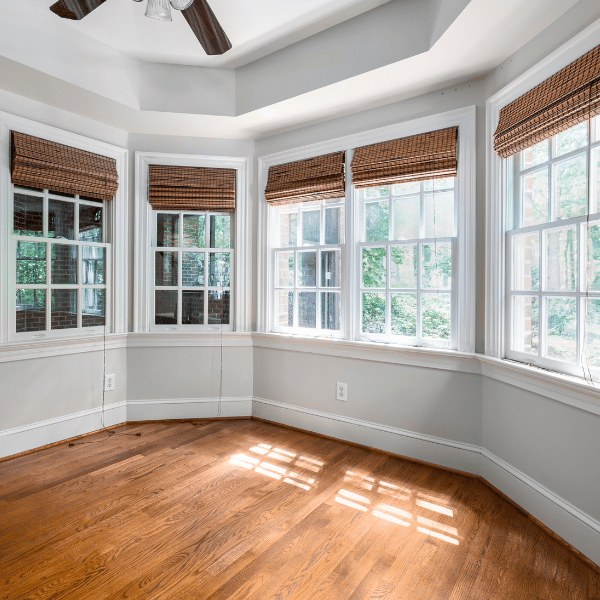 Bright, empty room with wood floors, white walls, multiple windows and doors featuring brown woven shades, and sunlight streaming in. Bright, empty room with wood floors, white walls, multiple windows and doors featuring brown woven shades, and sunlight streaming in.