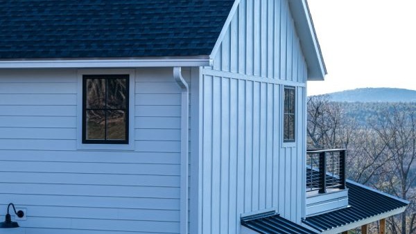 Side view of a modern white house with black-trimmed window, metal roof, outdoor lamp, and balcony, featuring stylish stucco siding—an elegant example of Utah new construction set against a backdrop of trees and distant hills.
