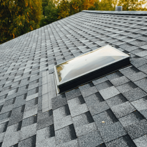 Gray asphalt shingle roof with a rectangular skylight; trees are visible in the background. Gray asphalt shingle roof with a rectangular skylight; trees are visible in the background.