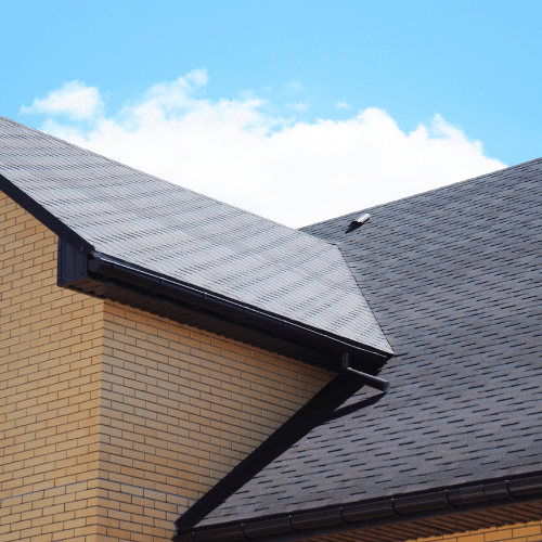 A close-up view of a house roof with dark gray shingles and yellow brick walls under a blue sky with some clouds. A close-up view of a house roof with dark gray shingles and yellow brick walls under a blue sky with some clouds.