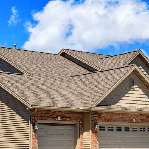 Suburban house exterior with tan siding, brick accents, two closed garage doors, and a multi-angled shingle roof under a partly cloudy sky. Suburban house exterior with tan siding, brick accents, two closed garage doors, and a multi-angled shingle roof under a partly cloudy sky.