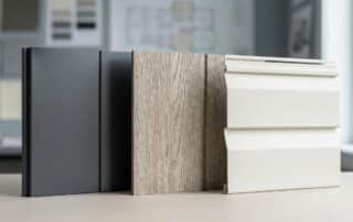 Three building material samples—dark gray fiber cement siding, wood-textured vinyl siding, and white metal siding—are displayed upright on a table in a well-lit room with blurred background plans.
