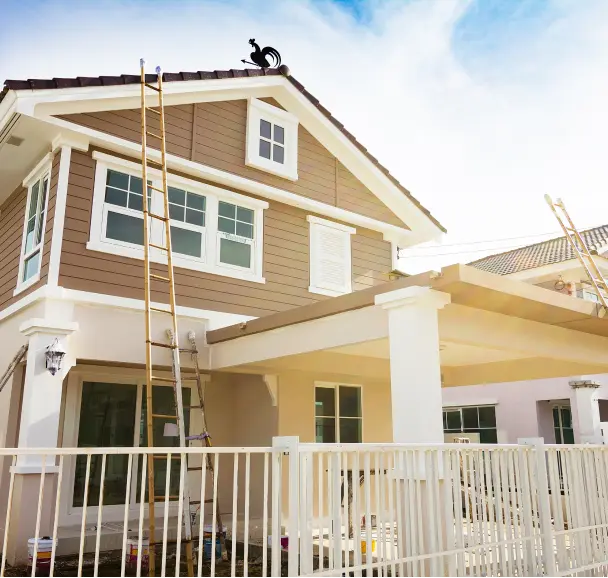 Two-story house with brown and white exterior, ladders leaning against the wall, and painting supplies, indicating ongoing siding remodeling work. Two-story house with brown and white exterior, ladders leaning against the wall, and painting supplies, indicating ongoing siding remodeling work.