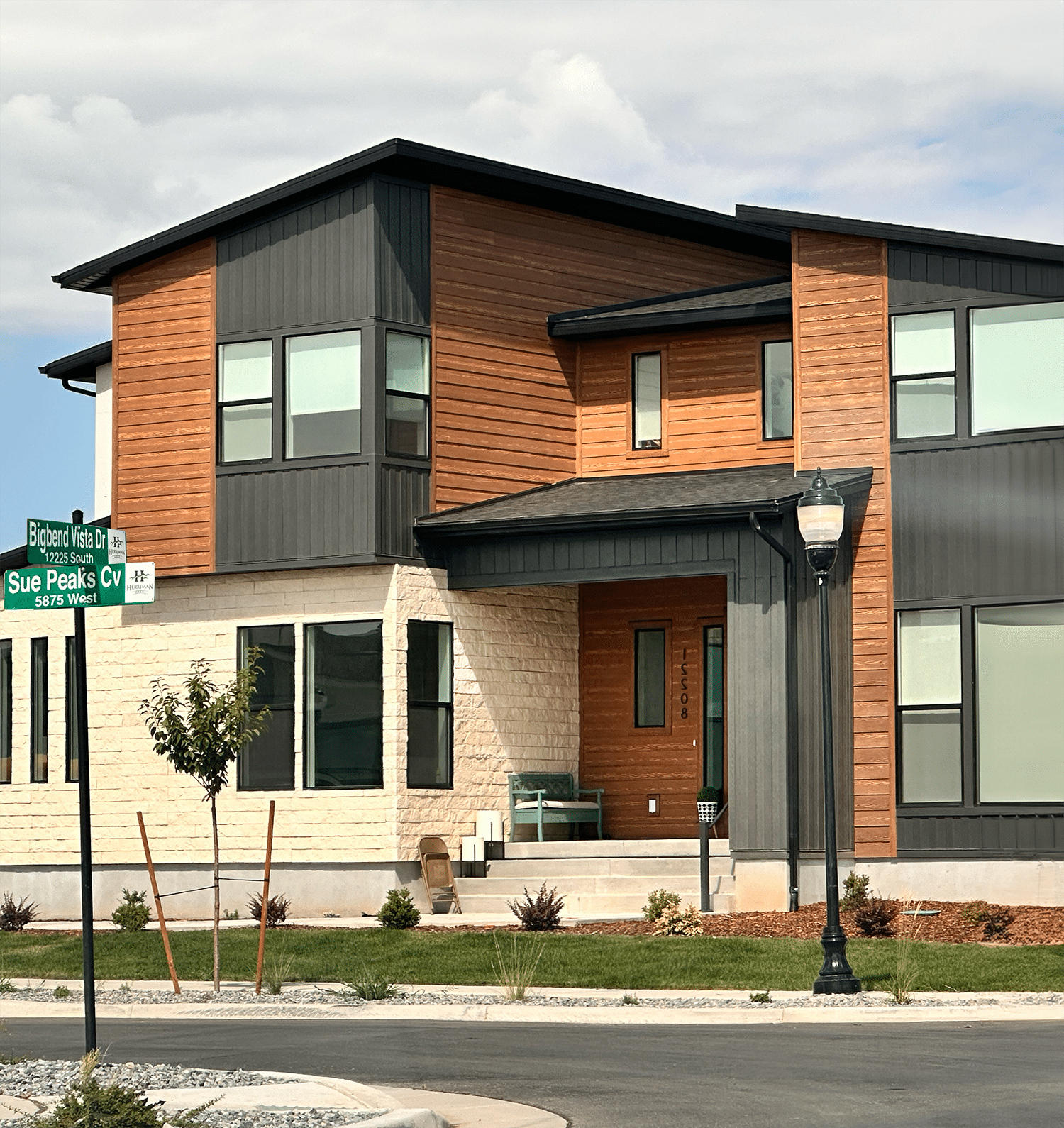Modern two-story house with mixed wood and dark panel exterior, large windows, and a small front porch. A street sign in the foreground reads “Bigbend Vista Dr” and “Sue Peaks Cv. Modern two-story house with mixed wood and dark panel exterior, large windows, and a small front porch. A street sign in the foreground reads "Bigbend Vista Dr" and "Sue Peaks Cv.