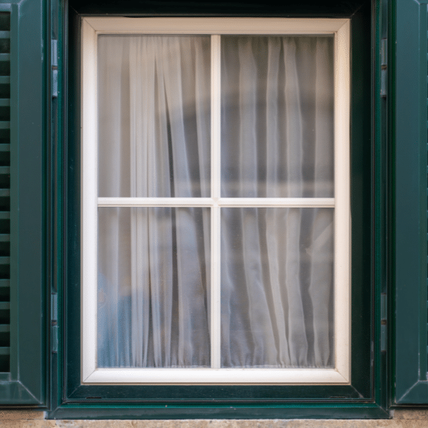 A closed window with white curtains behind glass panes, framed by green shutters and a green window frame—an inviting scene typical of Salt Lake City homes following expert vinyl window installation in Utah. A closed window with white curtains behind glass panes, framed by green shutters and a green window frame—an inviting scene typical of Salt Lake City homes following expert vinyl window installation in Utah.